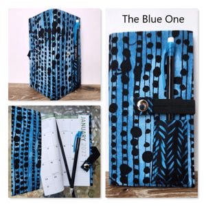 Op de afbeelding: Een stoffen plannerhoes met een abstract stippen- en strepenpatroon in blauw en zwart. De hoes bevat een zwarte elastische band met een zilveren metalen drukknoop en een klein vakje met een zwarte pen. De tekst The Blue One is zichtbaar.