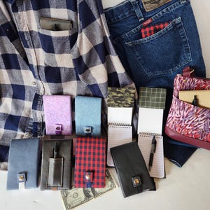 May include: An assortment of small notebooks and journals in various colors and patterns, including plaid, camouflage, and solid hues. A denim shirt and jeans are in the background, along with a patterned purse. A one-dollar bill is also visible.