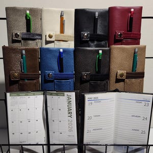 Op de afbeelding: Acht rechthoekige planners met gestructureerde covers in grijs, crème, zwart, rood, bruin, blauw, donkergrijs en beige, elk met een penhouder en drukknoopsluiting. Twee open planners tonen kalenderpagina's voor januari 2026 en mei 2025.
