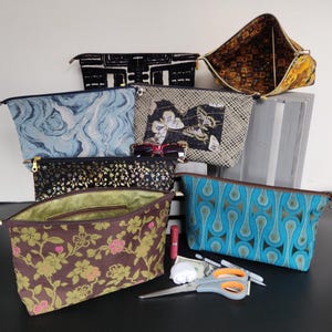 May include: A collection of zippered cosmetic bags in various patterns and colors. The bags include designs such as floral, geometric, and abstract patterns. Some bags are open, revealing the interior. Accessories like scissors, pens, and lipstick are also visible.