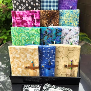 May include: A display of fabric-covered notebooks with various patterns, including floral, geometric, and abstract designs. Some notebooks have pen holders and leather accents. Two composition books with black and white speckled covers are in the foreground.