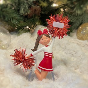 May include: A red and white cheerleader ornament with a red bow and pom-poms. The ornament has a blank white tag for personalization.
