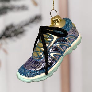 May include: A glass Christmas ornament shaped like a running shoe. The ornament is primarily blue with glitter accents, a black shoelace, and a gold-colored top. The sole is light blue. The ornament is suspended by a gold string.