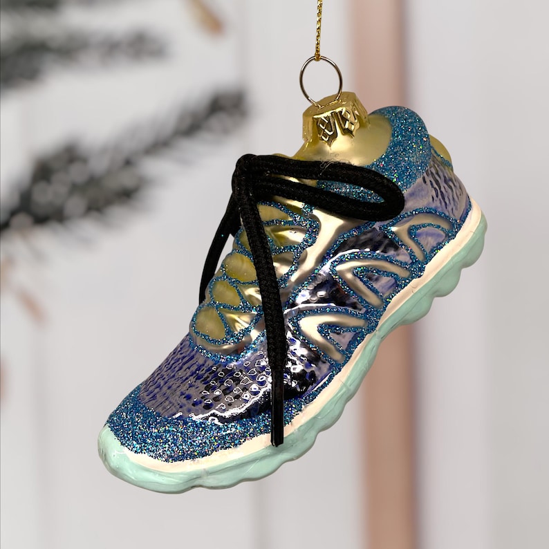 May include: A glass Christmas ornament shaped like a running shoe. The ornament is primarily blue with glitter accents, a black shoelace, and a gold-colored top. The sole is light blue. The ornament is suspended by a gold string.