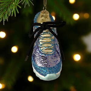 May include: A blue and silver glass Christmas ornament shaped like a running shoe. The ornament has black shoelaces and is decorated with glitter. The ornament is hanging from a gold-colored cap.