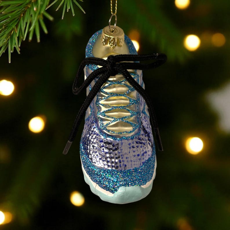 May include: A blue and silver glass Christmas ornament shaped like a running shoe. The ornament has black shoelaces and is decorated with glitter. The ornament is hanging from a gold-colored cap.