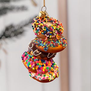Puede incluir: Una pila de cuatro adornos navideños con forma de rosquilla. Cada adorno es de un color diferente, incluyendo marrón, rosa y naranja, y está cubierto de chispas de colores. Los adornos están suspendidos por un hilo dorado.