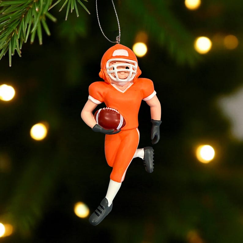 Peut inclure: Ornement de joueur de football am&eacute;ricain orange, avec un casque et des d&eacute;tails blancs. Le joueur tient un ballon de football marron et court. L'ornement est suspendu &agrave; un fil argent&eacute;, parfait pour un sapin de No&euml;l sur le th&egrave;me du sport.