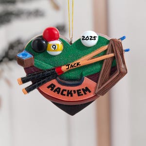 May include: A heart-shaped ornament featuring a miniature pool table scene. It includes a red 8-ball, a black ball, and a 9-ball. The ornament has two pool cues, a rack, and the text "RACK'EM" and "JACK". The year "2025" is also visible.