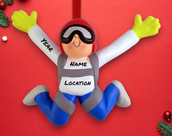 Può includere: Un ornamento da appendere a forma di paracadutista con un casco rosso, una tuta bianca e grigia e pantaloni blu. L'ornamento ha le parole "YEAR", "NAME" e "LOCATION" scritte su di esso.