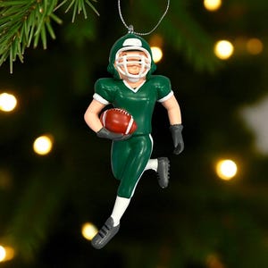 Peut inclure: Ornement de joueur de football am&eacute;ricain vert et blanc tenant un ballon de football marron. L'ornement porte un casque, un maillot et un pantalon verts. Le joueur court, avec une ficelle argent&eacute;e pour l'accrocher.