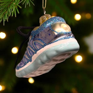 May include: A blue and silver glass Christmas ornament shaped like a sneaker. The ornament has a glittery blue trim and a black shoelace. The sneaker is hanging from a gold-colored metal cap.