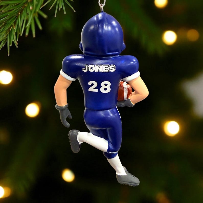 Peut inclure: Ornement de No&euml;l repr&eacute;sentant un joueur de football am&eacute;ricain en bleu. Le joueur porte le nom "JONES" et le num&eacute;ro "28" dans le dos. Il tient un ballon de football marron et porte un casque et un uniforme bleus. L'ornement est suspendu &agrave; un crochet argent&eacute;.