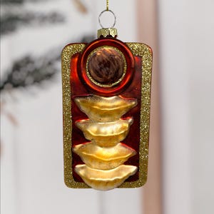 May include: A festive Christmas ornament shaped like a traffic light. The ornament features a red background with gold glitter trim. The lights are gold and the top light is a brown sphere. A gold loop is at the top for hanging.