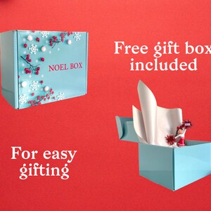 Puede incluir: Una caja de regalo azul con un dise&ntilde;o de copos de nieve blancos y bayas rojas. La caja dice "Noel Box" en la parte delantera. La caja est&aacute; abierta y tiene papel de seda blanco en el interior. El texto "Caja de regalo gratuita incluida" est&aacute; encima de la caja y el texto "Para facilitar el regalo" est&aacute; debajo de la caja.