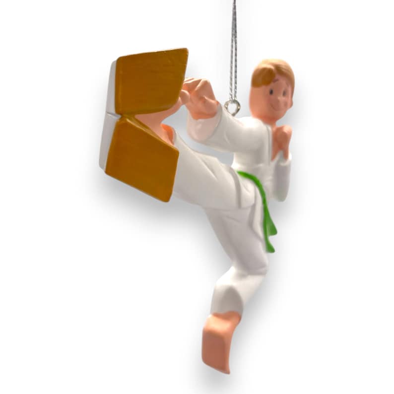 May include: A white karate ornament with a green belt, kicking a gold block. The ornament is designed to hang on a Christmas tree.