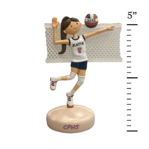 Puede incluir: Una figura de 12,7 cm de alto de una jugadora de voleibol con un uniforme blanco y azul con el nombre "Katie" y el n&uacute;mero "8" en la parte delantera. La jugadora est&aacute; rematando un bal&oacute;n de voleibol con el a&ntilde;o "2022" en &eacute;l. La figura est&aacute; de pie sobre una base con el texto "CPHS" en ella.