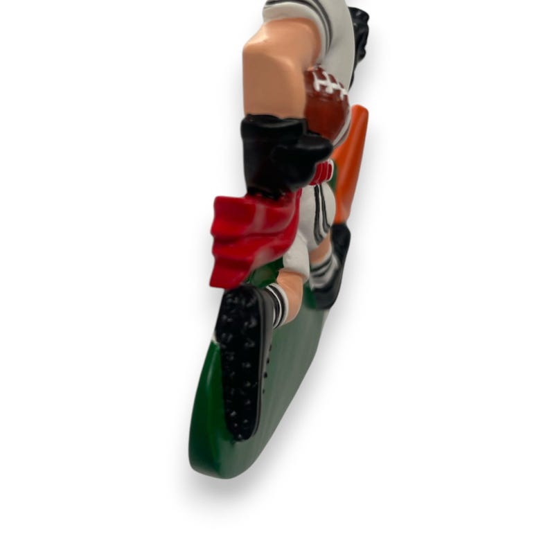 May include: A green and orange football field ornament with a white and black football player figurine holding a brown football. The player is wearing a red cape and is in a diving pose.