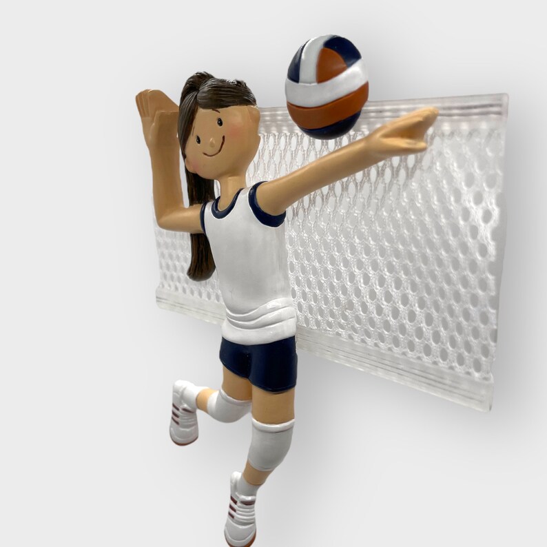 Puede incluir: Una figura de jugador de voleibol blanco y azul con cabello casta&ntilde;o, que lleva una camiseta blanca y pantalones cortos azules, est&aacute; rematando un bal&oacute;n de voleibol sobre una red blanca.