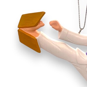 May include: A white figurine of a person in a karate pose, wearing a white gi and a purple belt. The figurine is holding a wooden board in their hand, which is broken in half.
