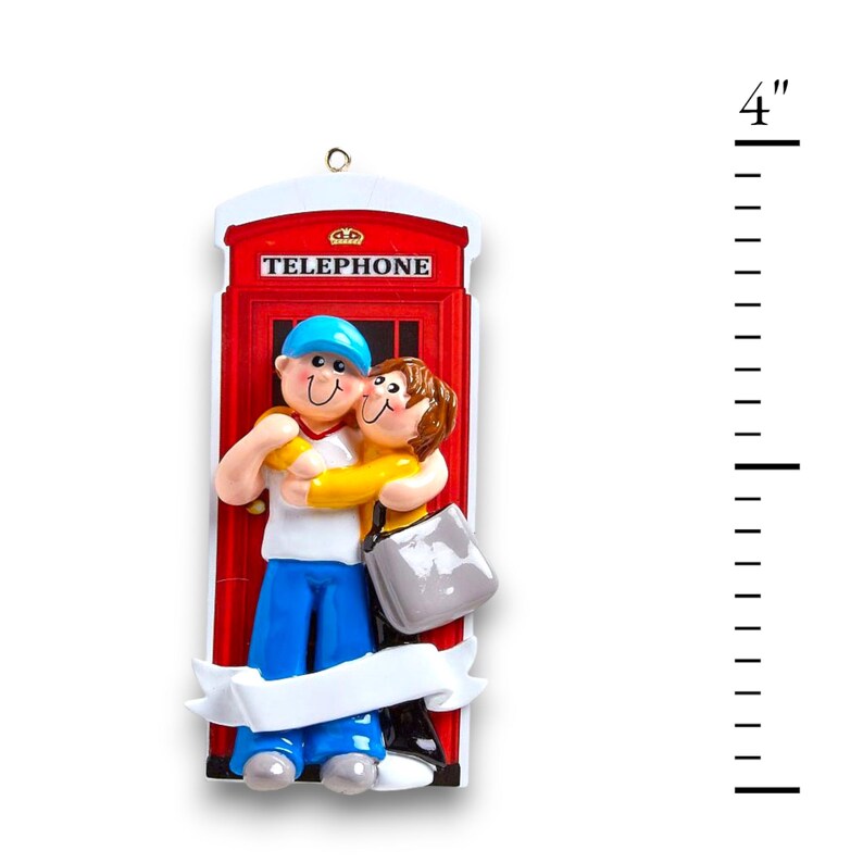 May include: A red telephone box Christmas ornament with a couple hugging inside. The ornament is 10 cm tall and has a white banner at the bottom.