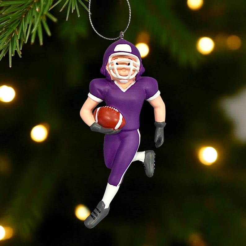 Peut inclure: Ornement de joueur de football am&eacute;ricain violet avec un casque et des bordures de maillot blancs. Le joueur court avec un ballon de football marron. L'ornement est suspendu &agrave; une ficelle argent&eacute;e, pour les f&ecirc;tes.