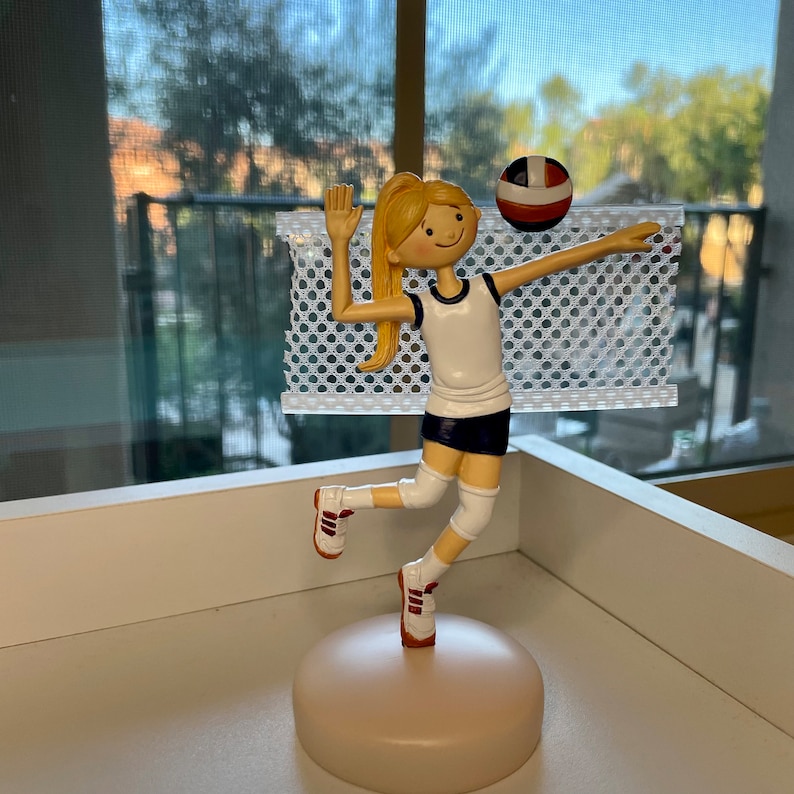 Puede incluir: Una figura de una jugadora de voleibol con cabello rubio, vistiendo una camiseta blanca, pantalones cortos azules y zapatillas rojas y blancas. La jugadora est&aacute; sosteniendo un bal&oacute;n de voleibol y est&aacute; saltando en el aire. La figura est&aacute; sobre una base beige y frente a una red blanca.