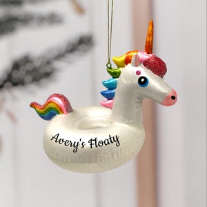Può includere: Ornamento a forma di unicorno bianco con criniera e coda arcobaleno. L'unicorno ha una finitura glitterata e un corno dorato. Il testo "Avery's Floaty" è scritto sul galleggiante. L'ornamento è appeso a un filo d'oro.