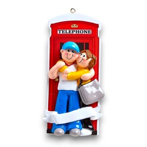 May include: A red telephone box Christmas ornament with a couple hugging in front of it. The man is wearing a blue hat and blue jeans. The woman is wearing a yellow shirt and black trousers. The ornament has a white banner at the bottom.