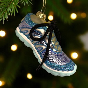 May include: A blue and silver glittery sneaker Christmas ornament with a black shoelace. The ornament is hanging from a gold-colored metal cap. The sneaker has a light blue sole and is hanging in front of a blurred background of Christmas lights and a green tree.