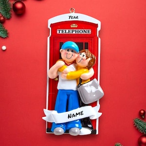 May include: A red telephone box Christmas ornament with a couple hugging in front of it. The ornament has the words "YEAR" and "TELEPHONE" on it. There is a banner at the bottom that says "NAME".
