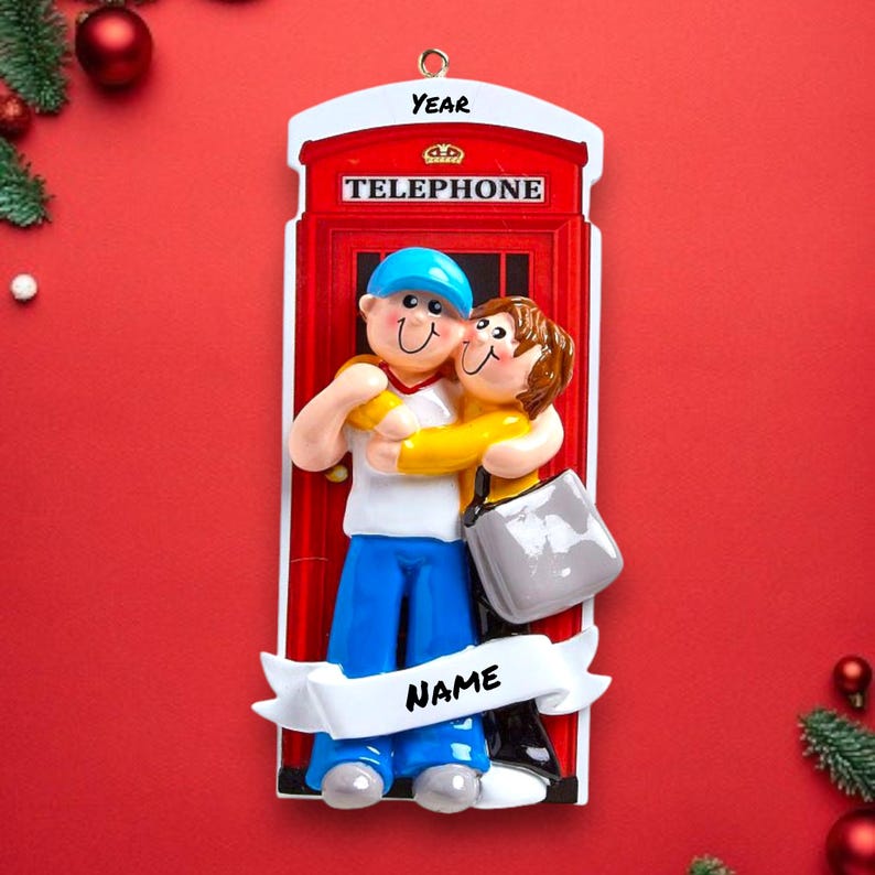 May include: A red telephone box Christmas ornament with a couple hugging in front of it. The ornament has the words "YEAR" and "TELEPHONE" on it. There is a banner at the bottom that says "NAME".