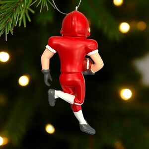 Peut inclure: Ornement de No&euml;l repr&eacute;sentant un joueur de football am&eacute;ricain en rouge, avec un casque et un uniforme. Il tient un ballon marron. D&eacute;tails blancs sur les manches et chaussettes, gants et chaussures gris.