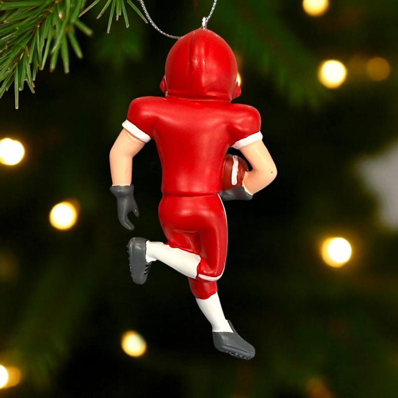 Peut inclure: Ornement de No&euml;l repr&eacute;sentant un joueur de football am&eacute;ricain en rouge, avec un casque et un uniforme. Il tient un ballon marron. D&eacute;tails blancs sur les manches et chaussettes, gants et chaussures gris.