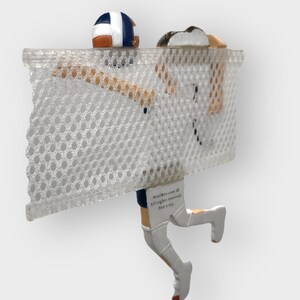 Puede incluir: Una figura blanca de un jugador de voleibol rematando un bal&oacute;n de voleibol. El jugador lleva un uniforme azul y blanco y calcetines blancos. El jugador est&aacute; saltando sobre una red blanca con un fondo blanco. La figura est&aacute; unida a una base blanca. El texto "NostalBox.com &copy; Todos los derechos reservados No es un juguete" est&aacute; impreso en la base.