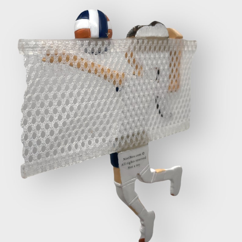 Puede incluir: Una figura blanca de un jugador de voleibol rematando un bal&oacute;n de voleibol. El jugador lleva un uniforme azul y blanco y calcetines blancos. El jugador est&aacute; saltando sobre una red blanca con un fondo blanco. La figura est&aacute; unida a una base blanca. El texto "NostalBox.com &copy; Todos los derechos reservados No es un juguete" est&aacute; impreso en la base.