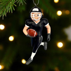 Peut inclure: Ornement de No&euml;l repr&eacute;sentant un joueur de football am&eacute;ricain en uniforme noir et blanc, tenant un ballon de football marron. Le joueur porte un casque. L'ornement est suspendu par un fil argent&eacute;.