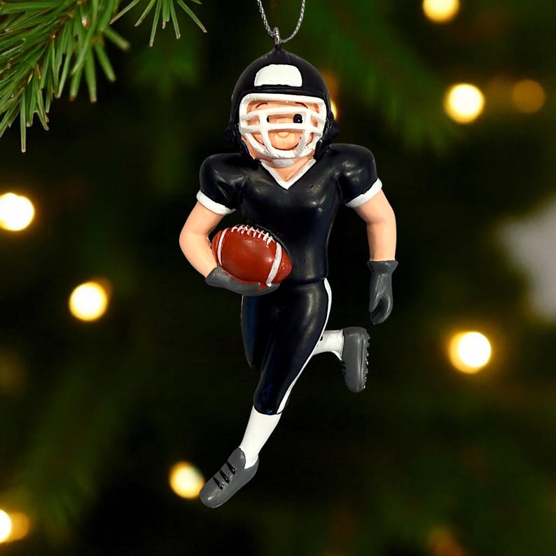Peut inclure: Ornement de No&euml;l repr&eacute;sentant un joueur de football am&eacute;ricain en uniforme noir et blanc, tenant un ballon de football marron. Le joueur porte un casque. L'ornement est suspendu par un fil argent&eacute;.
