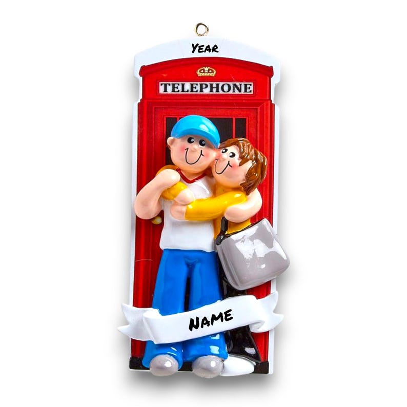 May include: A red telephone box Christmas ornament with a couple hugging in front of it. The ornament has a white banner with the word "NAME" on it and the words "YEAR" and "TELEPHONE" on the box.