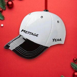Op de afbeelding: Witte baseballpet met een zwarte klep. Op de pet staat "MESSAGE" en "YEAR" geschreven. De pet hangt aan een rood touwtje.