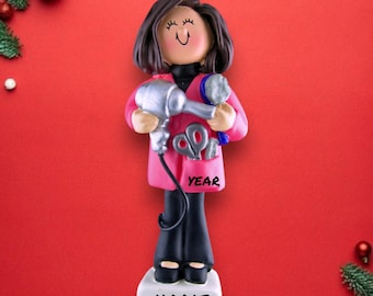 Op de afbeelding: Een roze en zwarte kerstversiering met een kapper die een föhn, schaar en kam vasthoudt. De versiering heeft de woorden "NAME" en "YEAR" erop.