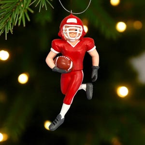 Peut inclure: Ornement de joueur de football am&eacute;ricain rouge, portant un casque blanc et tenant un ballon marron. Le joueur porte un uniforme rouge avec des bordures blanches, des gants et des chaussures noirs. L'ornement est suspendu &agrave; un fil argent&eacute;.