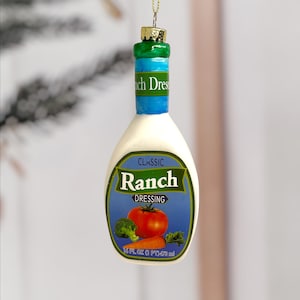Puede incluir: Un adorno navideño novedoso con forma de botella de aderezo ranch. La botella es blanca con una etiqueta azul y verde con un tomate, brócoli y zanahoria. La etiqueta dice "Classic Ranch Dressing". La parte superior es verde y azul.