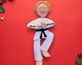 May include: A white karate ornament with a black belt and the words "YEAR" and "NAME" on the uniform. The ornament has a smiling face and is hanging on a red ribbon against a red background with Christmas decorations.