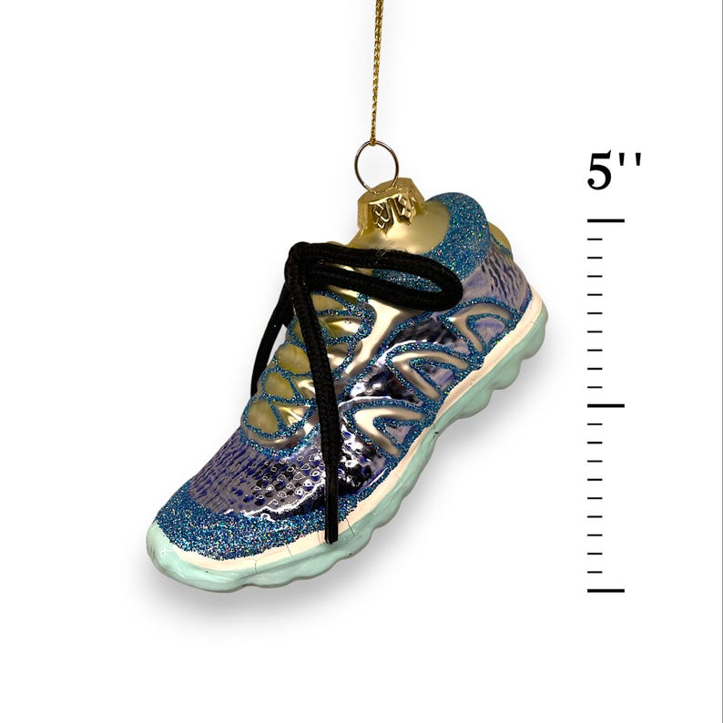 May include: A blue and silver glass ornament shaped like a running shoe, approximately 5 inches in length. The ornament has black laces and a gold-colored top. The shoe has a glittery blue trim and a light blue sole.