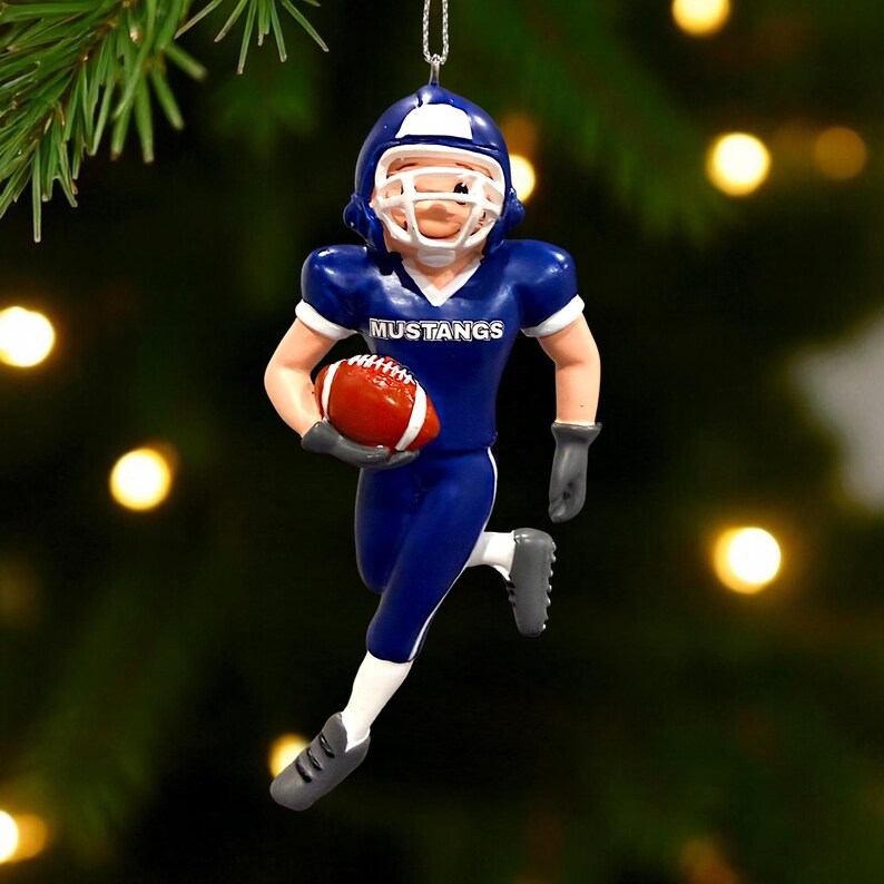 Peut inclure: Ornement de No&euml;l sur le th&egrave;me du football. L'ornement repr&eacute;sente un joueur de football en uniforme bleu avec le mot "Mustangs" sur la poitrine, tenant un ballon de football rouge et blanc. Le joueur porte un casque blanc et des gants et chaussures gris.
