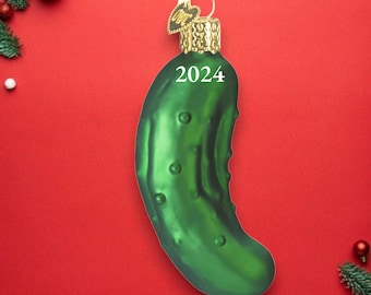 Può includere: Un ornamento di vetro verde a forma di cetriolo con una parte superiore dorata e l'anno 2024 scritto su di esso.