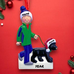 May include: A Christmas ornament featuring a person walking a black dog, both wearing Santa hats. The person is dressed in green and blue, and the dog is on a leash. The ornament has the word "NAME" on the hats and "YEAR" on the base.