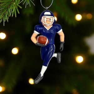 Peut inclure: Ornement de No&euml;l repr&eacute;sentant un joueur de football am&eacute;ricain en uniforme bleu marine, avec un casque et tenant un ballon de football marron. L'ornement a des accents blancs et est en mouvement. L'arri&egrave;re-plan est flou avec des lumi&egrave;res vertes et jaunes.