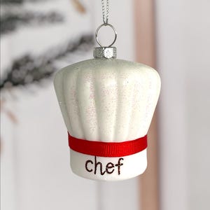 May include: A white chef hat ornament with a red band and the word "chef" in black script. The hat is covered in glitter and hangs from a silver chain, perfect for holiday decor.